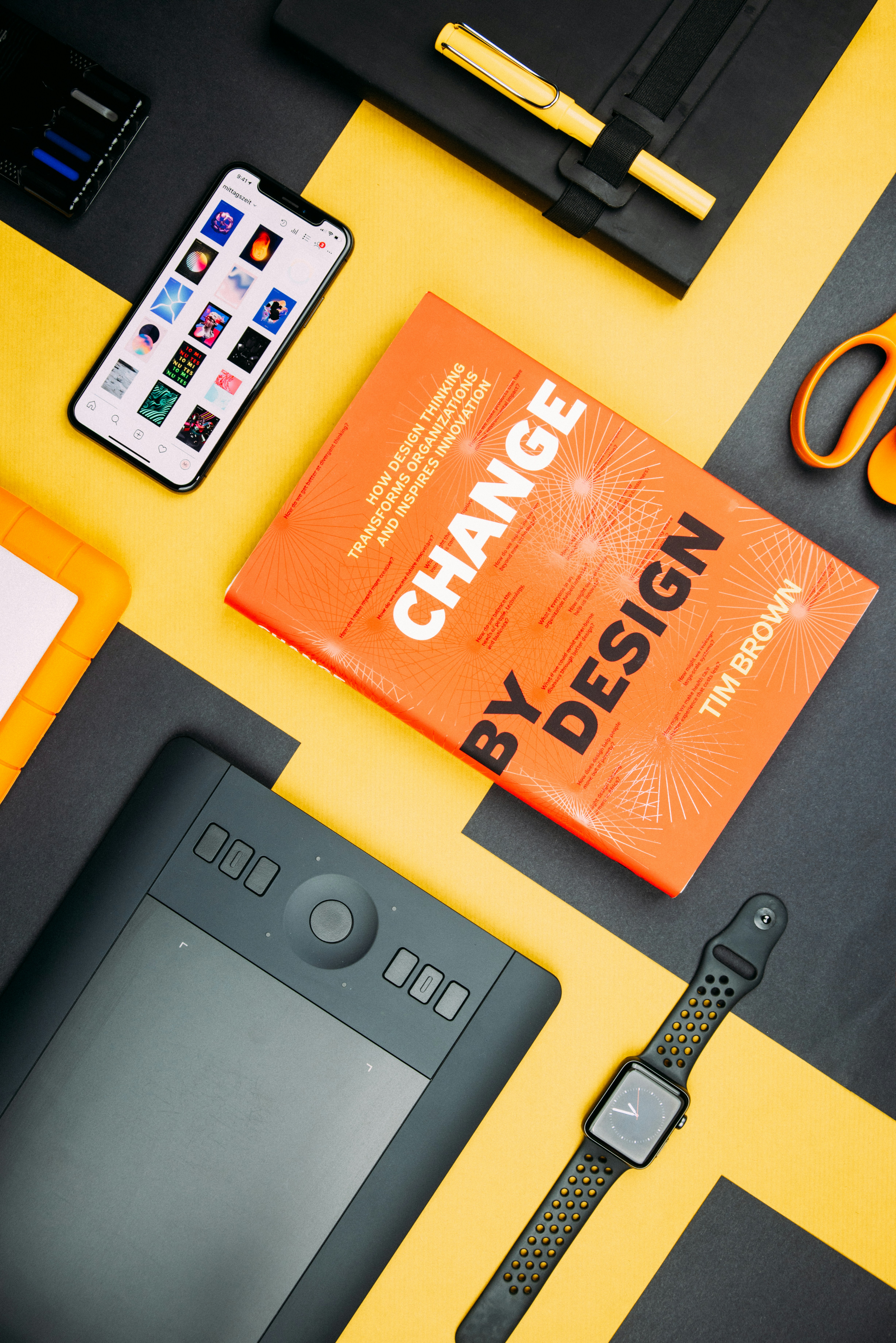 Overhead view of a creative workspace with a vibrant yellow and black color theme, featuring a book titled 'Change by Design' by Tim Brown, a smartphone displaying colorful graphics, a digital drawing tablet with stylus, a pair of scissors, and a smartwatch with a black and yellow band.